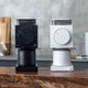 Two Fellow Ode Brew Grinder Gen 2 units—one black, one white—sit side by side on a wooden counter, each featuring Gen 2 Brew Burrs, a large front dial, and minimalist design with a blurred kitchen background.