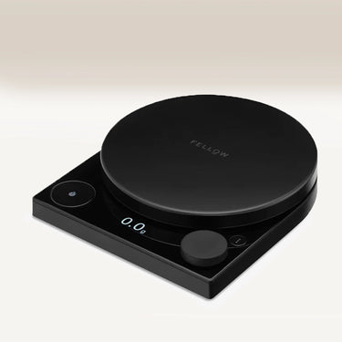 The Fellow Tally™ Pro Precision Scales by Fellow is a sleek black digital scale with a circular platform, LED display showing 0.0g, and control buttons—perfect for pour-over enthusiasts—shown against a light gradient background.
