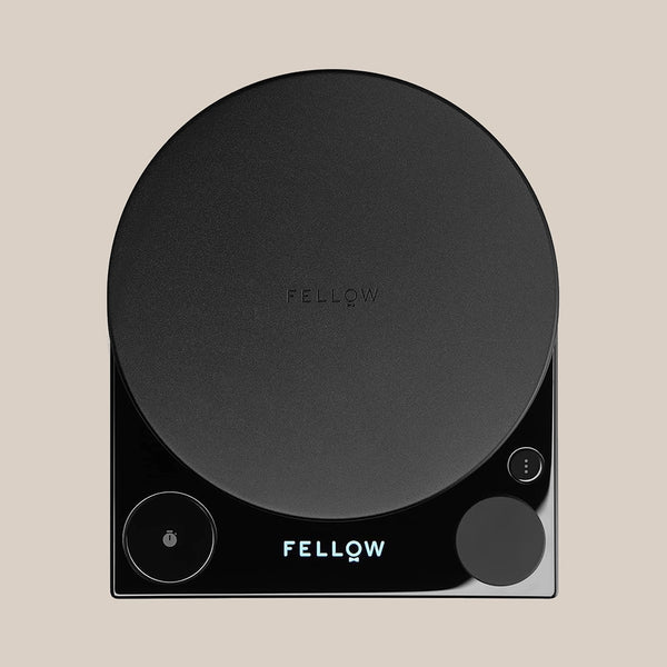 The Fellow Tally™ Pro Precision Scales by Fellow is a sleek black digital coffee scale with a round weighing platform, display, and two circular touch buttons—ideal for pour-over brewing—shown on a light beige background.