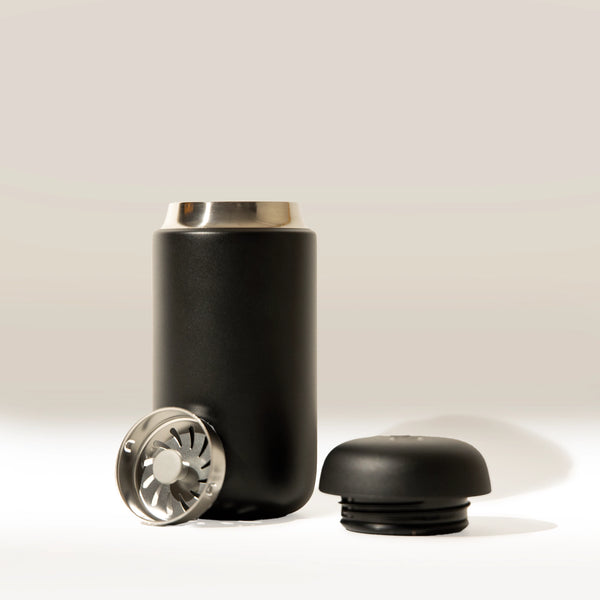 A Fellow Carter Move Mug (12oz) by Fellow in black stands upright with its stainless steel rim, while a matching black screw-on lid and round metal strainer are placed separately on a white surface against a pale backdrop.