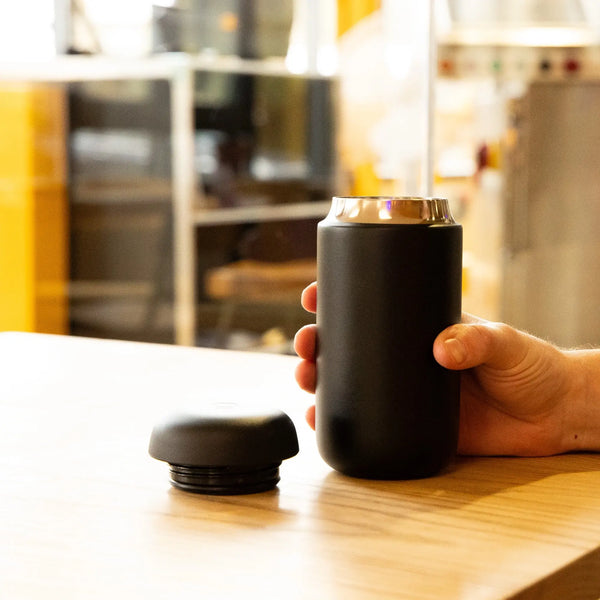 Someone holds the black, 12oz Fellow Carter Move Mug by Fellow, featuring a silver rim, on a wooden table. The lid sits nearby and shelves plus other equipment appear blurred in the indoor background.