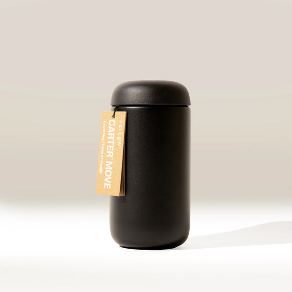 A tall, matte black Fellow Carter Move Mug (12oz) by Fellow with a rounded lid stands upright on a light surface. A brown tag reading "Carter Move" in white hangs from the vacuum insulated mug. The background is neutral and softly lit.