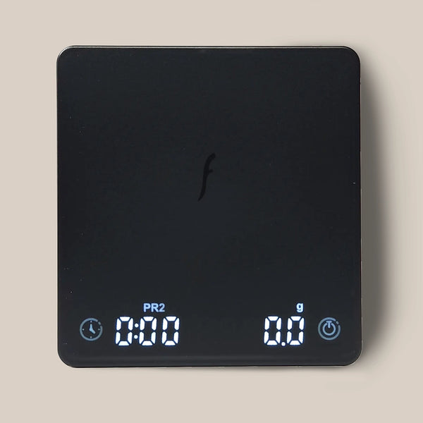 A black Flair Brew Scale by Flair, featuring a central "f" logo, displays 0:00 (left) and 0.0 g (right) on a light beige background—ideal as a precision coffee scale for espresso lovers.