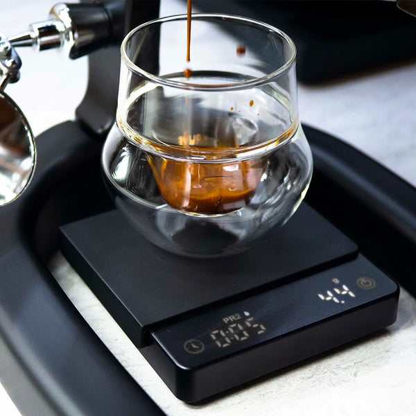 A shot of espresso is poured into a double-walled glass on the Flair Brew Scale by Flair, with the espresso machine and part of the scale’s display visible.