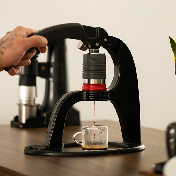 A hand presses down on the Flair NEO Flex Espresso Maker by Flair, extracting espresso into a glass on a wooden table. A flow-control portafilter, another coffee machine, and a plant blur in the background—a true home barista setup.