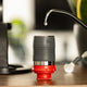 A close-up of the Flair NEO Flex Espresso Maker by Flair, in black and red, rests on a wooden surface as water drips from its spout. The blurred background with green and black objects highlights a home barista’s setup.