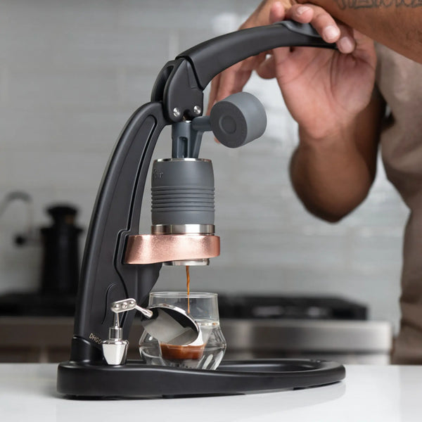 Using a Flair PRO 3 Espresso Maker with a bottomless portafilter, someone brews coffee into a small glass cup on the kitchen counter, with a cozy background featuring a blurred stove and kettle.