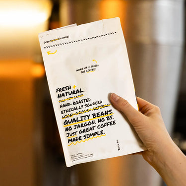 A hand holds a white bag labeled “Indian Monsoon Malabar Coffee Nº 53” in bold black and yellow text, describing it as fresh, hand-roasted, ethically sourced, natural, and premium—set against a softly blurred indoor background.