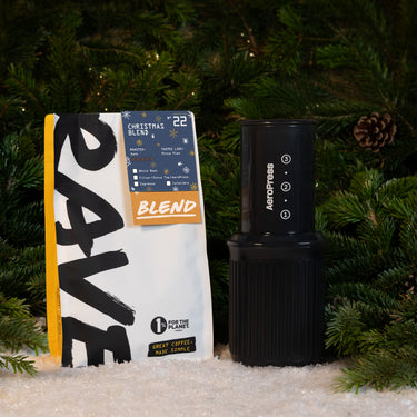An AeroPress Go Gift Set with a bag of Christmas Blend coffee beans sitting on a snowy surface in front of pine branches.