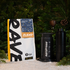 An AeroPress Gift Set with Grinder and a bag of Christmas Blend coffee on a snowy surface in front of pine branches.