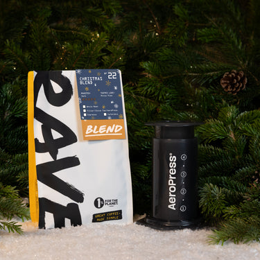 A bag of gourmet Christmas blend coffee next to an Aeropress Gift Set, set in front of pine branches and artificial snow.