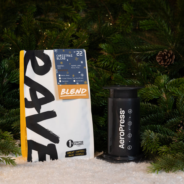 A bag of gourmet Christmas blend coffee next to an Aeropress Gift Set, set in front of pine branches and artificial snow.
