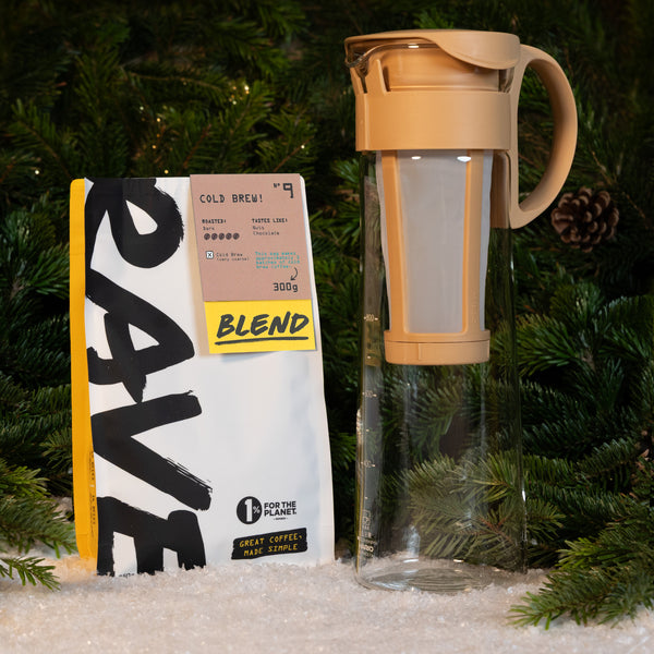 A Hario Cold Brew Gift Set featuring a coffee bag and a cold brew pitcher with a removable mesh filter, set against a background of evergreen branches and artificial snow.