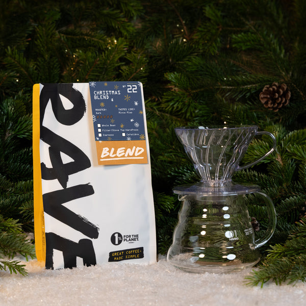 A Hario Hand Brewed Coffee Gift Set featuring a bag of Christmas blend coffee and a V60 pour-over dripper on a glass server, set against a background of evergreen branches and artificial snow.