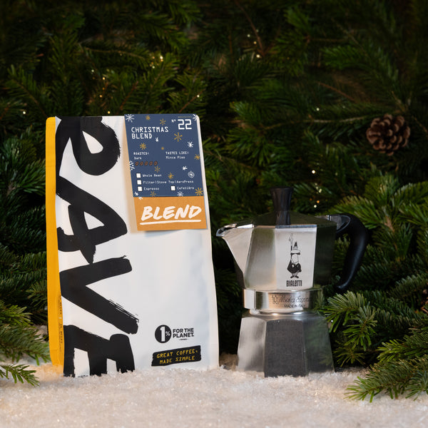 A bag of Christmas Blend coffee next to a Bialetti Moka Express Stove Top Gift Set, set against a background of evergreen branches and snow.