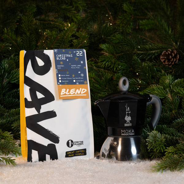 A bag of Rave Coffee Christmas blend next to a black Bialetti Induction Stove Top Gift Set on a bed of artificial snow in front of pine branches.
