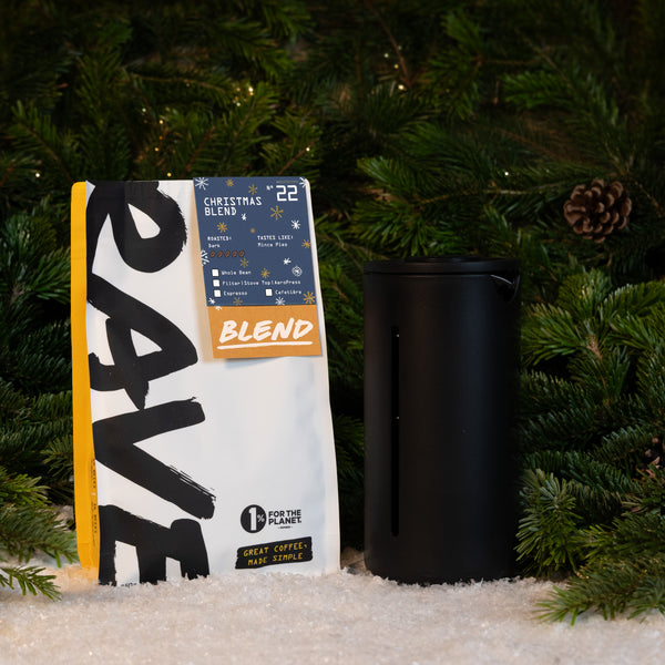 A bag of freshly roasted Christmas Blend coffee next to a black cylindrical Timemore French Press Gift Set on a bed of artificial snow in front of evergreen branches.