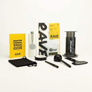 The AeroPress Gift Set with Grinder includes a yellow brew guide, fresh Rave coffee beans, manual grinder, AeroPress coffee maker, scoop, stirrer, filter holder, filters, and a black pouch—all displayed on a white background.