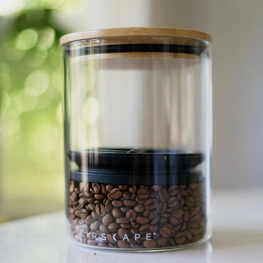 A Planetary Design Glass Airscape Coffee Storage Container with oxygen-removing Airscape technology holds brown coffee beans. It sits on a white surface against a blurred green background.