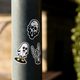 Showcased on a pole, the RAVE COFFEE Glow in the Dark Sticker Pack features three bold designs: a melting ghost face, a cartoon ghost with orange eyes and shoes, and a skeletal hand flashing the “rock on” sign. Each eye-catching sticker includes glow-in-the-dark accents that come alive after dark. Set against a blurred backdrop of sunlight and greenery, these playful stickers are perfect for fans of rave coffee culture.
