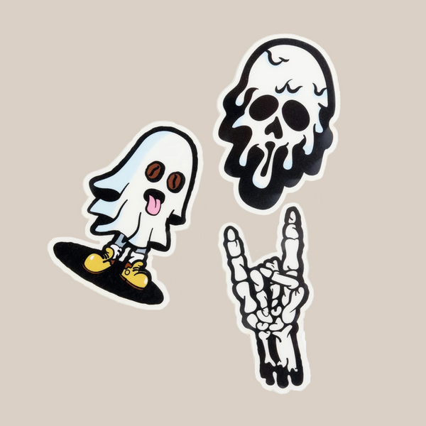The RAVE COFFEE Glow in the Dark Sticker Pack features three unique designs—a smiling ghost with yellow shoes, a melting skull, and a skeleton hand making the rock gesture—each glowing vividly against the plain beige background.