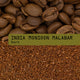 A close-up of coffee beans above coarsely ground Indian Monsoon Malabar Coffee Nº 53, with a green label in the center reading INDIA MONSOON MALABAR Dark in bold, typewriter-style font.
