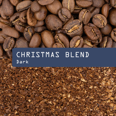 Whole roasted coffee beans of Christmas Blend Nº 22 rest above a blue rectangle with the text Christmas Blend and Dark over a background of rich, sweet ground coffee.