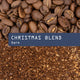 Whole roasted coffee beans of Christmas Blend Nº 22 rest above a blue rectangle with the text Christmas Blend and Dark over a background of rich, sweet ground coffee.