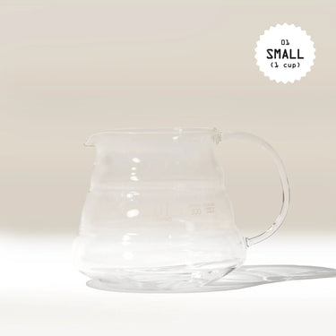 A clear Hario V60 Glass Coffee Server by Hario with a handle rests on a light surface, casting a soft shadow. Made from heat-proof glass, it pairs perfectly with V60 drippers. 