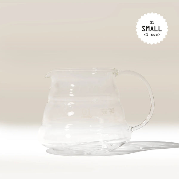 A clear Hario V60 Glass Coffee Server by Hario with a handle rests on a light surface, casting a soft shadow. Made from heat-proof glass, it pairs perfectly with V60 drippers. "01 SMALL (1 cup)" is shown in the top right corner.