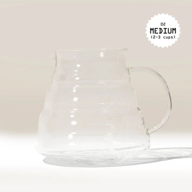 A Hario V60 Glass Coffee Server, made from heat-proof glass with a handle and measurement lines, sits on a white surface. In the top right corner, black text in a starburst reads 02 MEDIUM (2-3 cups)—perfect for V60 drippers.