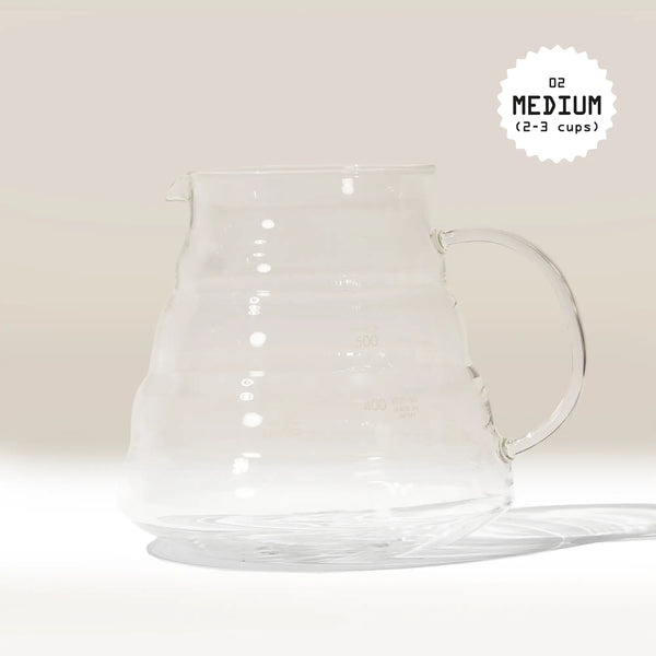 A Hario V60 Glass Coffee Server, made from heat-proof glass with a handle and measurement lines, sits on a white surface. In the top right corner, black text in a starburst reads 02 MEDIUM (2-3 cups)—perfect for V60 drippers.