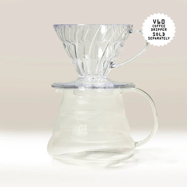 A Hario V60 Glass Coffee Server by Hario, made of heat-proof glass, is shown on a light background with its handle and spout visible. A transparent V60 dripper rests on top; note that V60 drippers are sold separately.