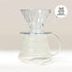 A Hario V60 Glass Coffee Server by Hario, made of heat-proof glass, is shown on a light background with its handle and spout visible. A transparent V60 dripper rests on top; note that V60 drippers are sold separately.