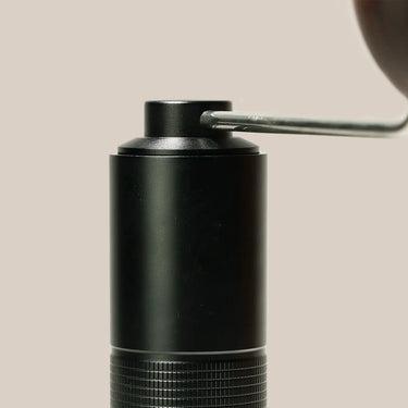 Close-up of the Hario Kabuto Coffee Grinder by Hario—a black metal cylinder with textured grip, metal handle, and stainless steel burr—set against a plain beige background.