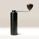The Hario Kabuto Coffee Grinder by Hario is a black, entry-level grinder with a metal handle, dark wooden knob, textured grip, and durable stainless steel burr, shown standing upright on a light background.