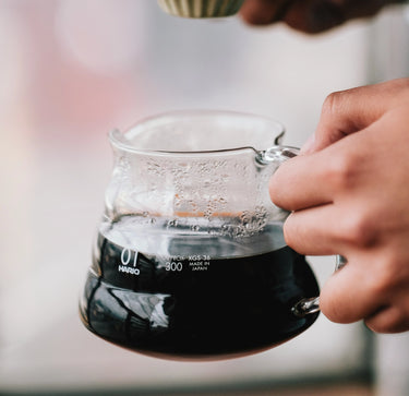 A close-up of a hand holding the Hario V60 Glass Coffee Server by Hario, filled with hot black coffee and steam rising from its heat-proof glass. This server, compatible with the V60 dripper, keeps your brew perfectly hot and aromatic.