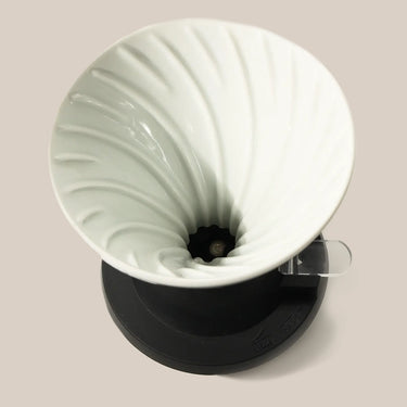 Top view of the Hario Switch V60 Ceramic Immersion Dripper (Size 02) by Hario, featuring spiral ridges on a black base and clear handle, set against a light beige background.
