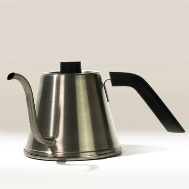The Hario Tetsu Kasuya Pour Over Kettle by Hario, made of stainless steel with a black handle and slim curved spout, sits on a light surface against a neutral background—ideal for precise pour over coffee brewing.