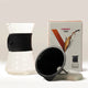 The Hario V60 Drip Decanter by Hario, featuring a borosilicate glass carafe, black silicone band, plastic dripper, and branded box, is displayed on a white surface.