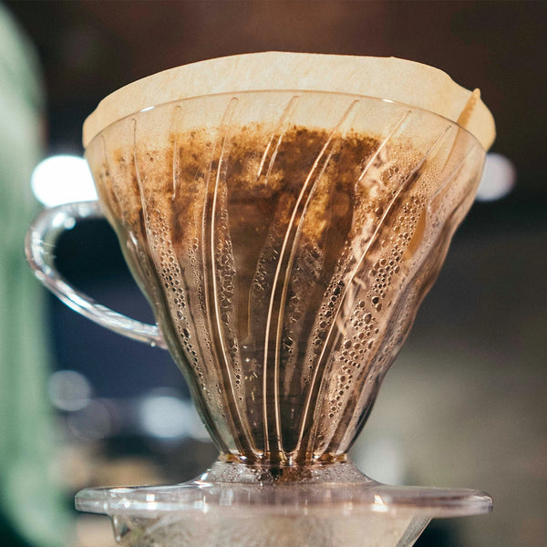 A close-up of the Hario V60 Clear Plastic Coffee Dripper Size 02 by Hario, featuring a paper filter with brewed coffee grounds and condensation on the clear dripper.