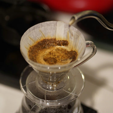 Hot water pours from a gooseneck kettle over ground coffee in a paper filter set in the Hario V60 Plastic Clear Coffee Dripper Size 01 by Hario, placed above a carafe.