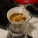 Hot water pours from a gooseneck kettle over ground coffee in a paper filter set in the Hario V60 Plastic Clear Coffee Dripper Size 01 by Hario, placed above a carafe.