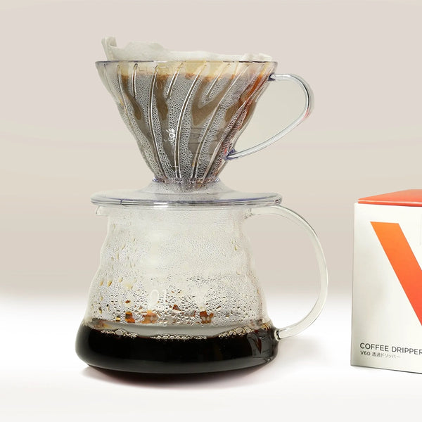 A Hario V60 Glass Coffee Server 01 by Hario with a pour-over dripper and paper filter sits on a heat-proof carafe, filled with fresh coffee. A box labeled “Coffee Dripper” is partially visible on the right.