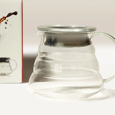 A Hario V60 Glass Coffee Server 01 with lid and handle by Hario rests on a white surface. On the left, part of its box displays the same heat-resistant glass server and a coffee splash illustration.