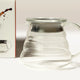 A Hario V60 Glass Coffee Server 01 with lid and handle by Hario rests on a white surface. On the left, part of its box displays the same heat-resistant glass server and a coffee splash illustration.