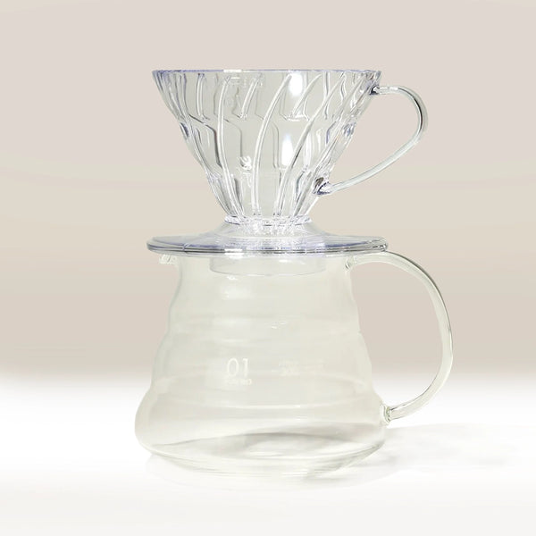 A clear Hario V60 pour-over dripper rests on a matching Hario V60 Glass Coffee Server 01, both with ribbed designs and handles from Hario, set against a light neutral background.