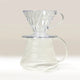 A clear Hario V60 pour-over dripper rests on a matching Hario V60 Glass Coffee Server 01, both with ribbed designs and handles from Hario, set against a light neutral background.