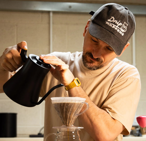Man pouring coffee from a black pitcher into a glass filter in a kitchen.