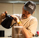Man pouring coffee from a black pitcher into a glass filter in a kitchen.